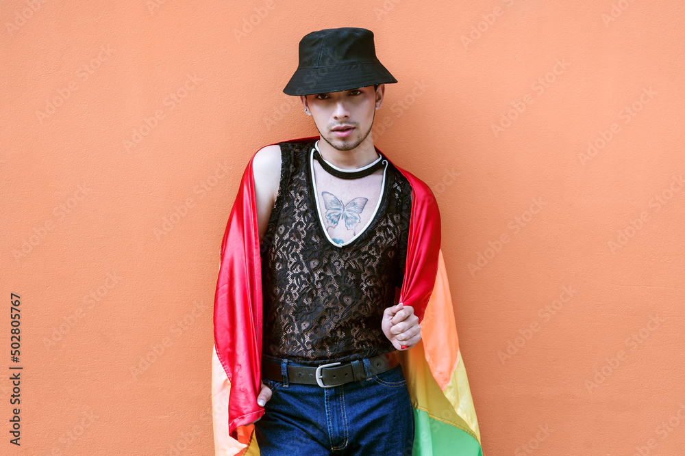 Handsome young man in lgbt community hat draped with gay flag on a coral colored wall.
