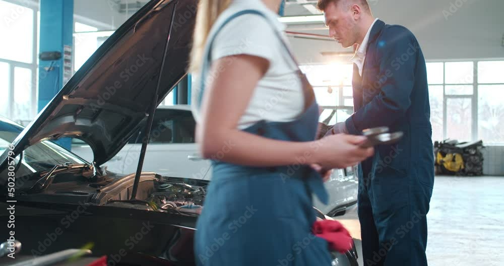 Caucasian male mechanic in uniform coming to car to repair motor in auto service and checking with light. Young man engineer fixing engine. Maintaining concept. Repairment work of technician.
