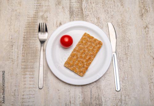 A single tomato and a slice of crispbread on a plain white plate and cutlery with signs of use on a rustic wooden surface
