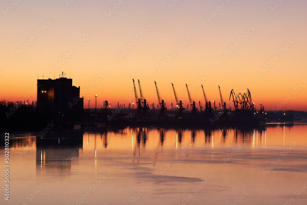 River landscape with silhouette of cranes in cargo port at sunrise on quiet surface of autumn Danube river