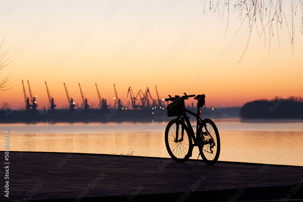 Fototapeta premium Silhouette of a bicycle against the backdrop of port cranes at dawn