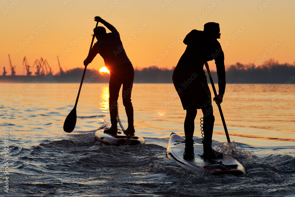 Silhouettes of a two men rowing on stand up paddle boarding (SUP) at sunrise on quiet surface of autumn Danube river. Morning training on SUP