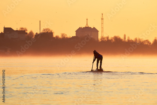 Silhouette of boy rowing on supboards (SUP) at calm river at orange sunset. Rear view on paddle boy