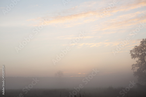 Beautiful foggy sunrise over misty field an early morning. Winter autumn countryside landscape. Fog down in valley, sun and fluffy clouds above horizon