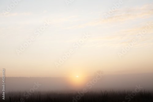 Beautiful foggy sunrise over misty field an early morning. Winter autumn countryside landscape. Fog down in valley, sun and fluffy clouds above horizon