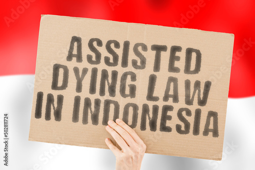 The phrase " Assisted dying law in Indonesia " on a banner in men's hands with a blurred Indonesian flag in the background. Pain. Care. Clinic. Sickness. Ethical. Ethic. Depression. Drug. End. Judge
