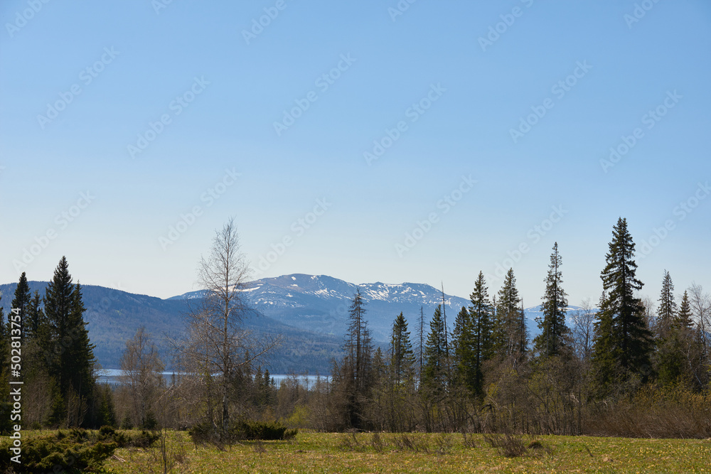 Scenic landscape of coniferous woods, lake and mountains covered with snow in the distance on a sunny day. National park, Ural region, Russia