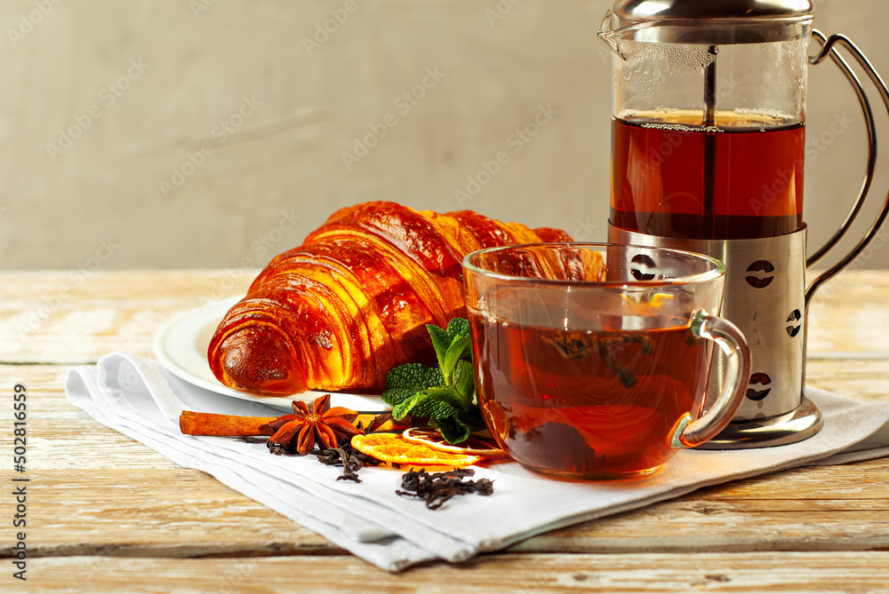 Croissant in a white plate with tea. Mug with tea on an old wooden table. Whole black tea, lemon, mint leaves and cinnamon on a napkin. Copy space and free space for text near food. Croissant in a white plate with tea. Mug with tea on an old wooden table. Whole black tea, lemon, mint leaves and cinnamon on a napkin. Copy space and free space for text near food.