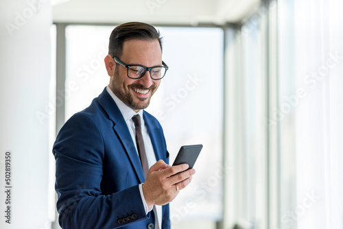 Handsome businessman wearing eyeglasses using smart phone and smiling while standing near the office window, Copy Space