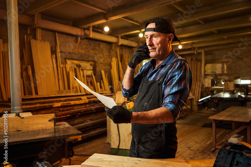 Portrait of a senior carpenter looking at blueprints plans and talking phone with a client in the carpentry workshop © bondvit