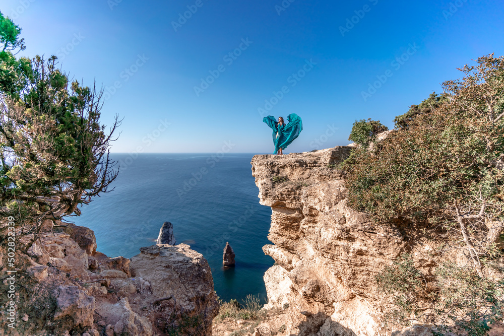 A girl with loose hair in a long mint dress descends the stairs between the yellow rocks overlooking the sea. A rock can be seen in the sea. Sunny path on the sea from the rising sun
