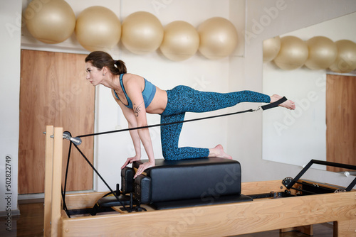 A young woman in sportswear does Pilates on a reformer, lifting her leg up © GRON777