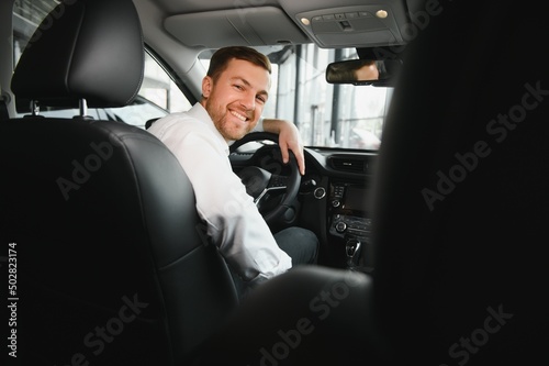 Taxi driver smiling while turning over to the backseat looking talking with clients. Successful businessman husband looking at the backseat at dealer shop before buying a car