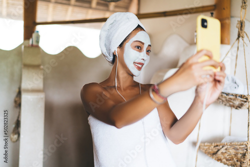 Smiling female in headphones using front mobile camera for clicking selfie images during time for doing beauty procedures for skin care, happy Asian woman in alginate mask enjoying dayspa in home photo