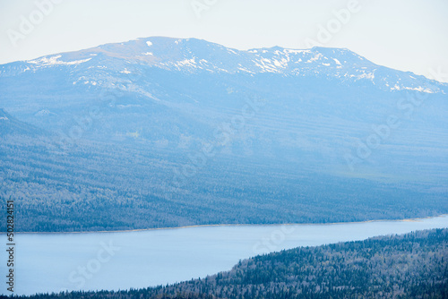 Scenic aerial view of vast woodland, high snow-capped mountains in the distance and wide river running through national park of the Urals, Russia