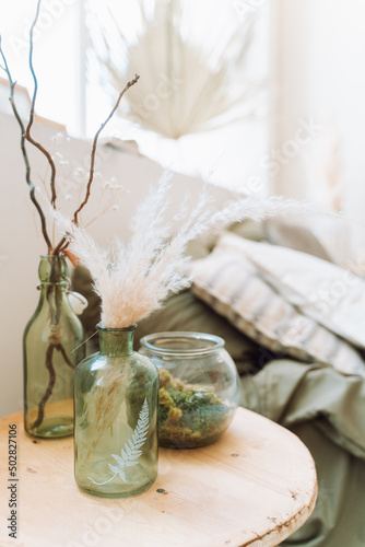 Interior of sunny modern bedroom with bed covered with grey bed linen. Different green glass bottles with dry decorative pampas, twigs, round glass vase with moss on beige round wooden bedside table. 