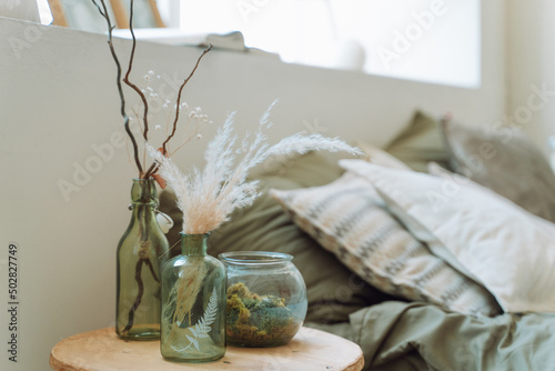 Interior of bright modern bedroom with bed covered with grey bed linen. Different green glass bottles with dry pampas, twigs, decorative plant, round glass vase with moss on beige bedside table.  photo