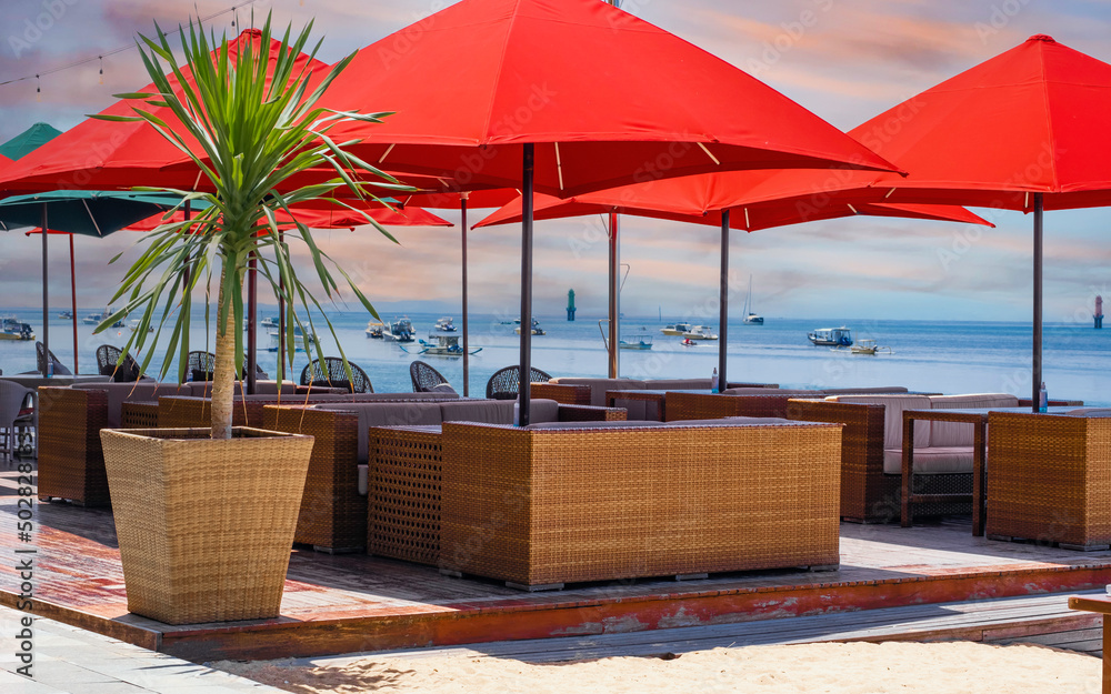 Obraz premium Sea side cafe with wicker furniture and bright red canvas umbrellas and a potted palm tree. Beautiful view of the ocean with boats scattered in distance. Early morning pink and blue sky. No people