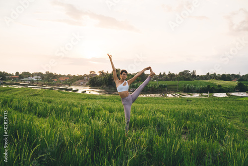 Flexible female motivated with sportive goals feeling wellness and vitality during summer yoga practice at rice fields, Caucasian woman in tracksuit stretching leg training body flexibility outdoors