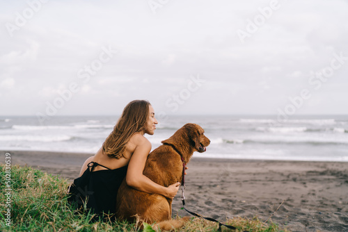 Carefree Caucasian woman in casual sundress clothing hug favorite dogs feeling love emotions during daily recreation at coastline, travel girl take care of cute pet enjoying together weekend #502830539