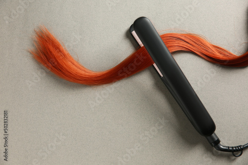Modern straightener with red hair lock on grey background, top view