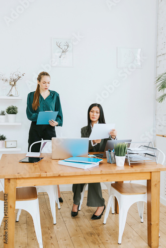 two young business women professionals in formal wear clothes work in modern office using laptop, tablet, brainstorm and search for solutions together, confident independent Asian girl solves problems