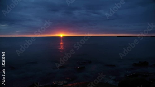 Time lapse of dramatic orange sunset over seascape ocean with clouds moving fast in dark blue sky and stones in water. Composition with long exposures