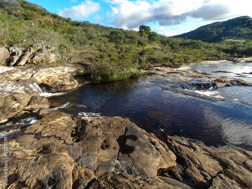 Pequeno curso d'água entre lindas pedras, com muita vegetação em volta e céu azul e nuvens, localizada na região de Três Barras, município do Serro, Minas Gerais, Brasil.