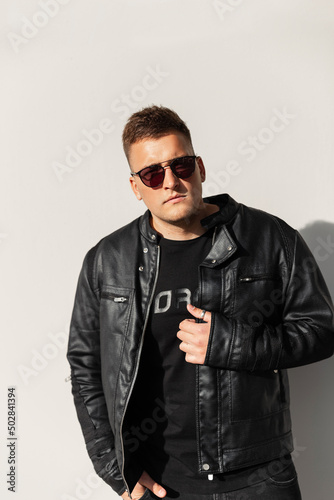 Handsome young man hipster model with a leather jacket with a black t-shirt and sunglasses stands near a wall on a sunny day