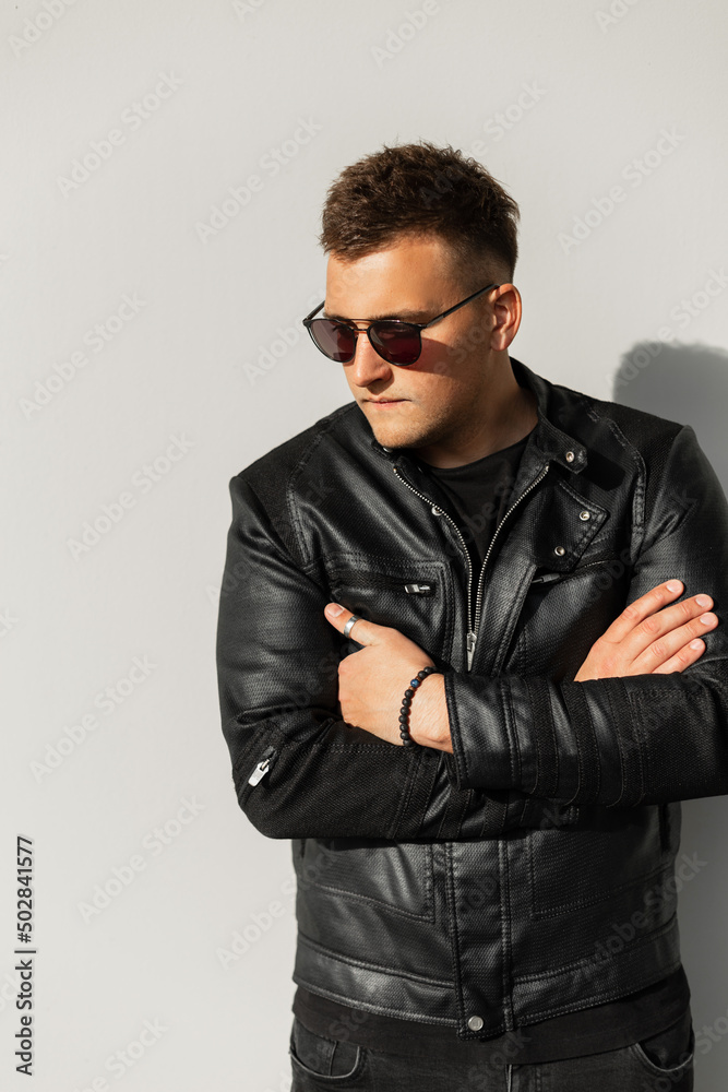 Plakat Stylish young hipster man model with hairstyle in black leather jacket with cool sunglasses stands near gray wall