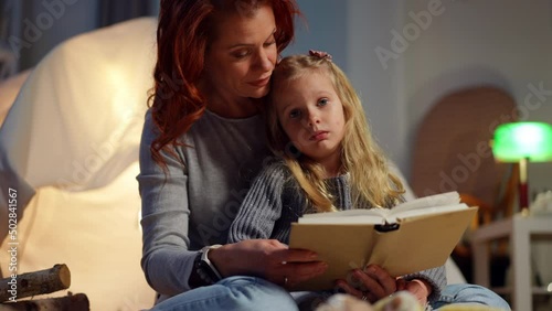 Portrait of cute girl listening to woman reading fairytale at bedtime at tent in living room. Calm Caucasian daughter enjoying leisure with mother at home at night. Family and childhood concept photo