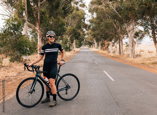 Female cyclist in sportswear wearing sunglasses and helmet relaxing after ride standing on empty road