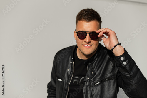 Happy handsome young man with hair in a fancy leather black jacket looks through fashion sunglasses at the camera and smiles against the gray concrete background