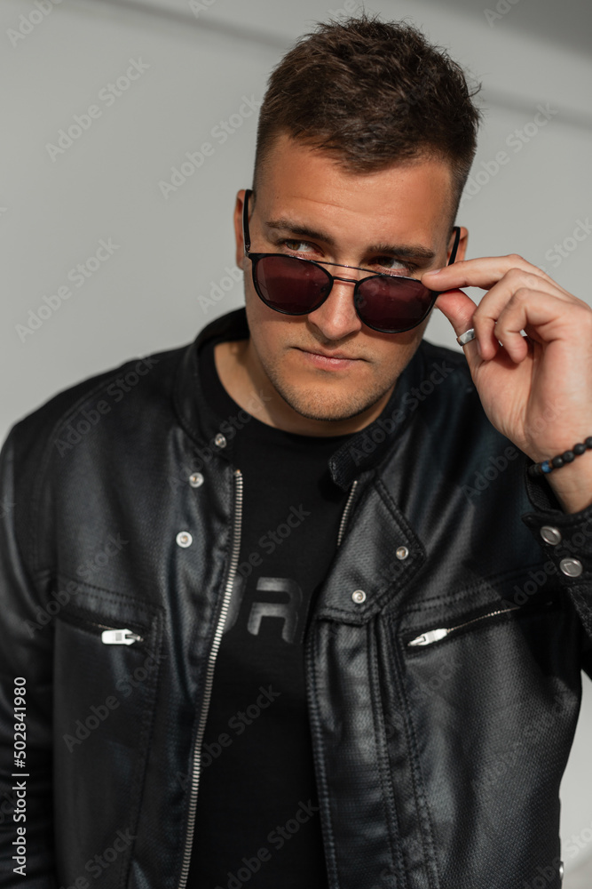 Handsome fashionable man in fashion black clothes with a leather jacket looks through sunglasses in the city Handsome fashionable man in fashion black clothes with a leather jacket looks through sunglasses in the city
