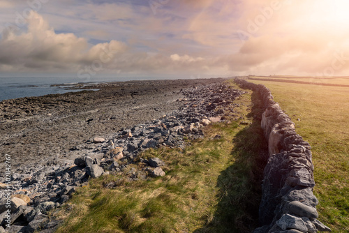 Long stone fence alongside coast line. Cloudy sky with sun flare in the background. Aran island, county Galway, Ireland. Irish nature landscape. photo