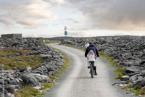 Family with children cycling on small narrow country road in rough stone terrain to a lighthouse. cloudy sky. Inisheer, Aran island, county Galway, Ireland. Irish landscape. Adventure concept. photo