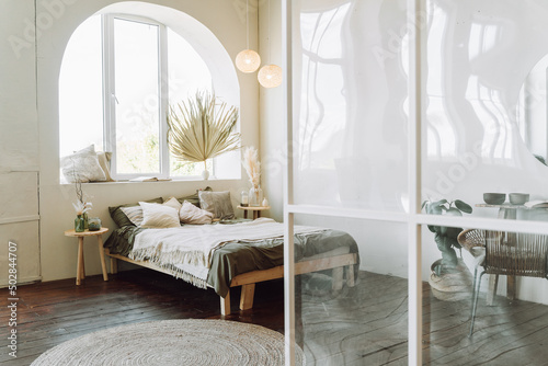 Interior of sunny modern bedroom with transparent plastic room divider . Wooden bed covered with grey bed linen, beige cotton plaid with fringe on brown floor. Different glass vases on bedside tables. photo