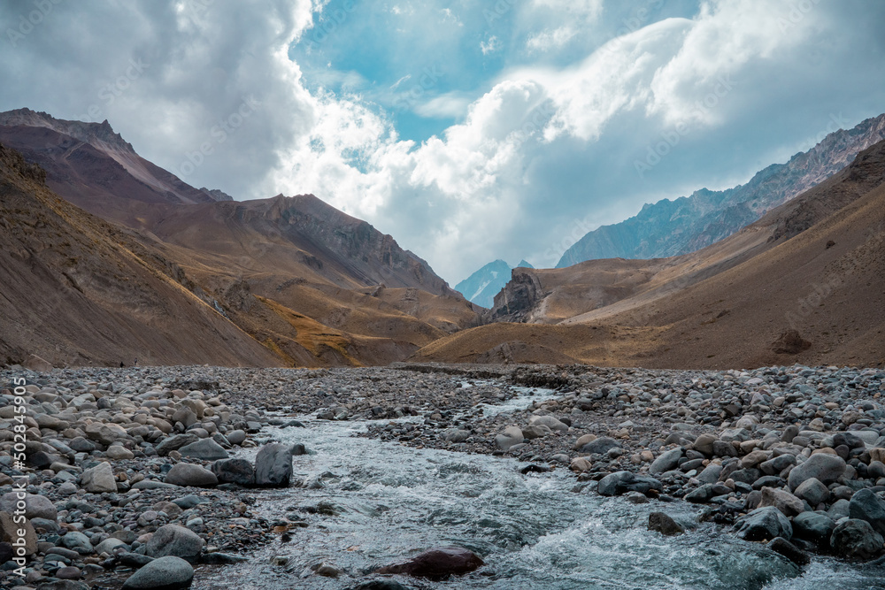 Horizontal shot of rushing river in rocky valley in Cajon del Maipo with clouds and mountains in the background, Chile