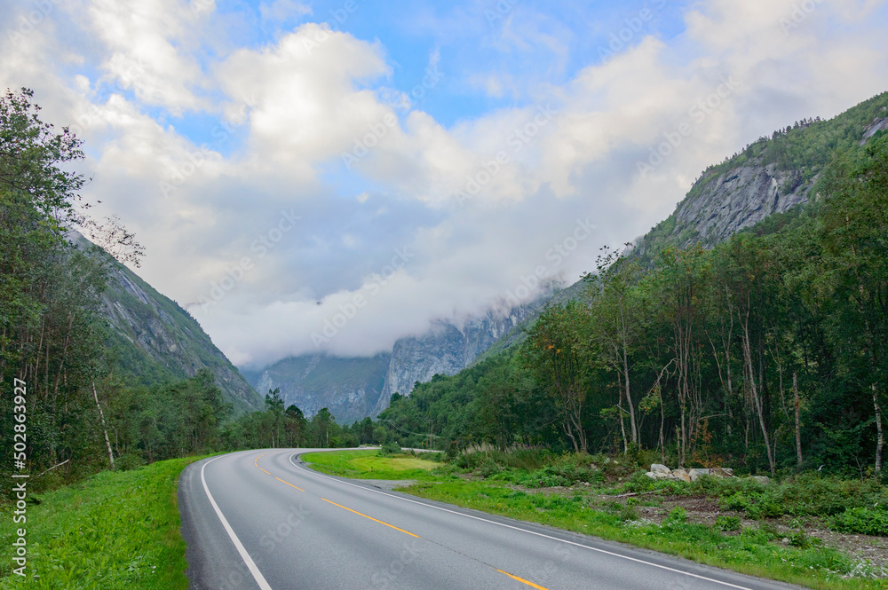 road to the mountains in Norway. Blue sky with clouds, grey asphalt, green trees and grass, mountain slopes far away.