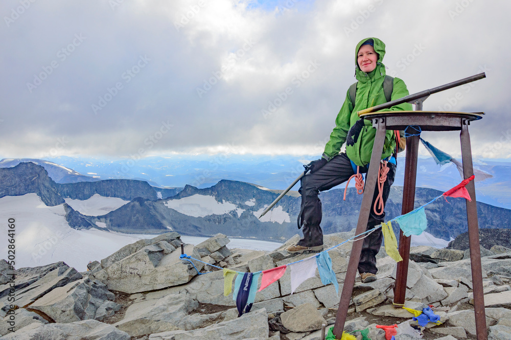 young woman alpinist standing on the top of the Galdhopiggen mountain summit in Norway. White and grey snow and ice, blue sky with clouds, rocks and dark mountains around