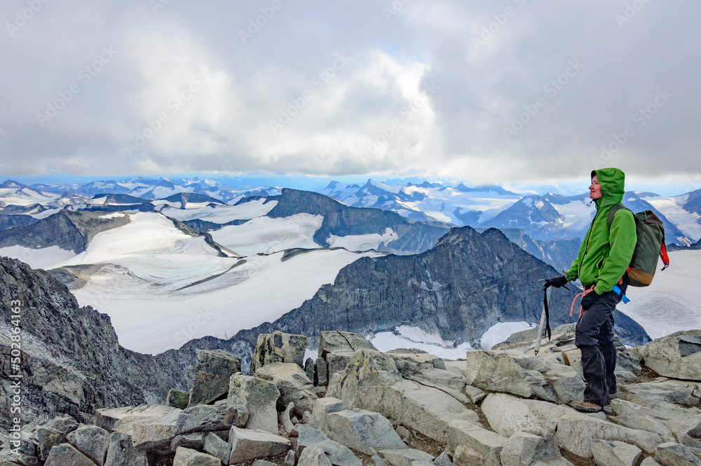 young woman climber standing on the top of the Galdhopiggen mountain summit in Norway. White and grey snow and ice, blue sky with clouds, rocks and dark mountains around