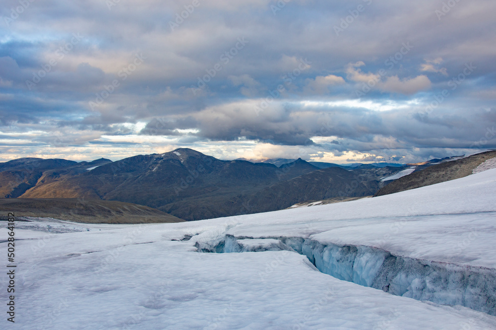 Evening on the glacier on the trail from Galdhopiggen summit in Norway. White, blue and grey cracked ice, dark mountains, sunset sky with clouds Evening on the glacier on the trail from Galdhopiggen summit in Norway. White, blue and grey cracked ice, dark mountains, sunset sky with clouds