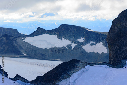 View of the mountain landscape in Jotunheimen national park in Norway from the Galdhopiggen mountain. Blue sky with clouds, grey and biege rocks, mountain slopes, white snow and ice of the glacier