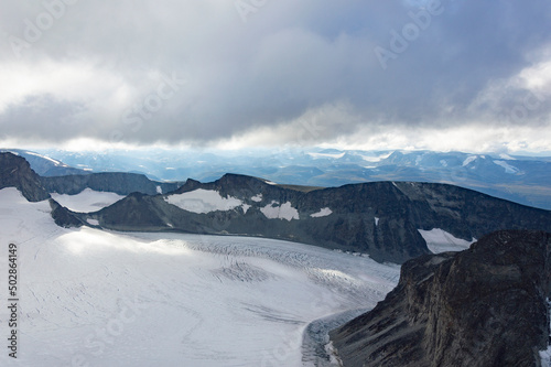 View of the mountain landscape in Jotunheimen national park in Norway from the Galdhopiggen mountain. Blue sky with clouds, grey and biege rocks, mountain slopes, white snow and ice of the glacier
