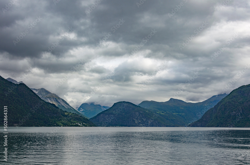 View of Geirangerfjord in Norway. Grey sea water, cloudy sky, dark green and blue mounain slopes View of Geirangerfjord in Norway. Grey sea water, cloudy sky, dark green and blue mounain slopes