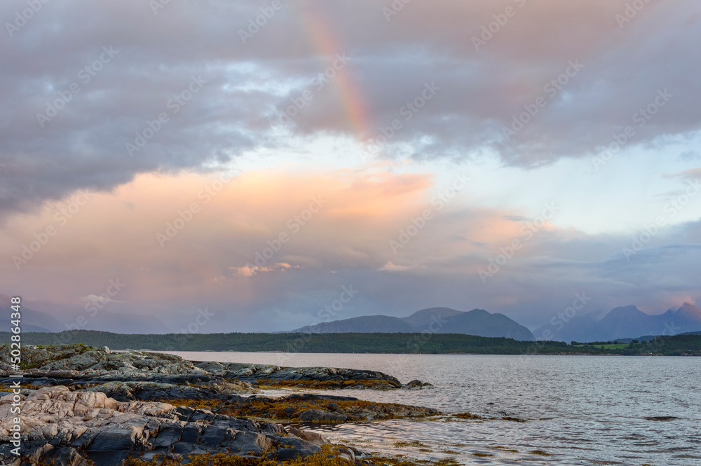 Norwegian landscape: blue evening sky with clouds  above the fjord, rainbow, calm water and misty mountains far away
