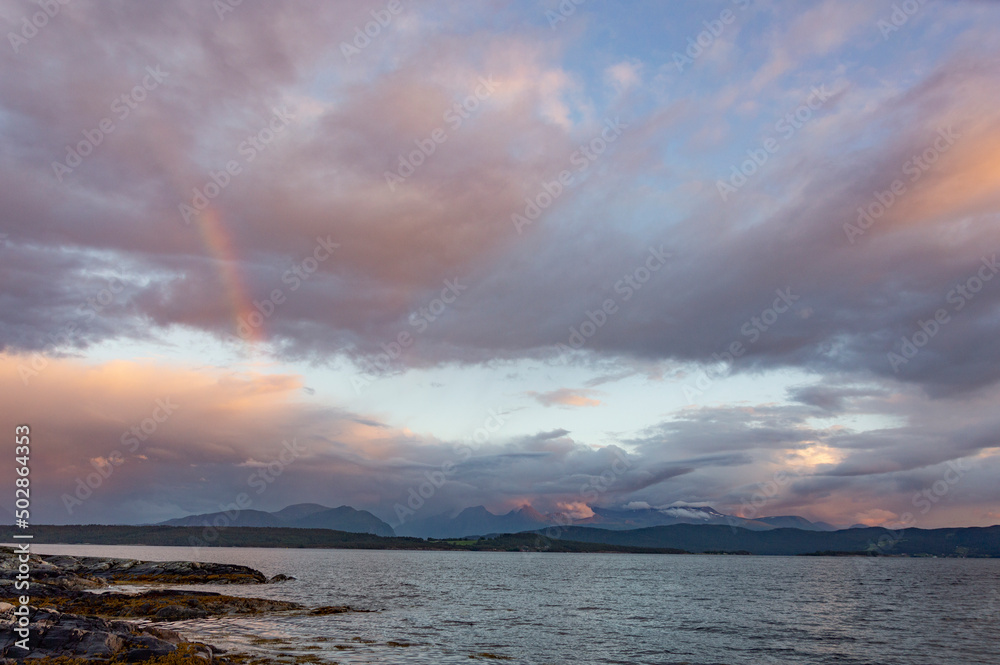 Norwegian landscape: blue evening sky with clouds  above the fjord, rainbow, calm water and dark mountains far away