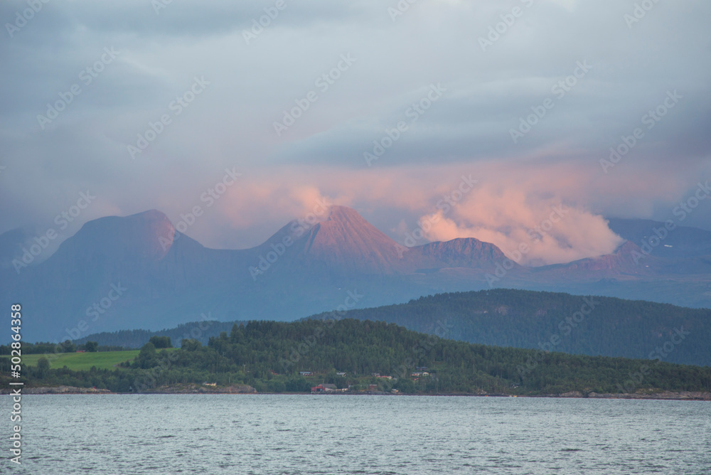 Sunset landscape in Norway. Blue and grey sea water, green shore, misty mountains on the background