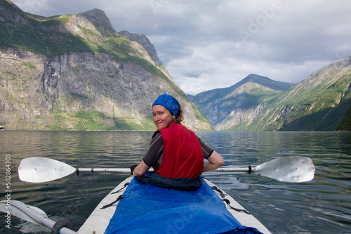 Young woman paddling in Geirangerfjord in Norway. Blue and grey sea water, blue sky with clouds, green and mounain slopes with sunshine and sadows on the forest, white and blue canoe