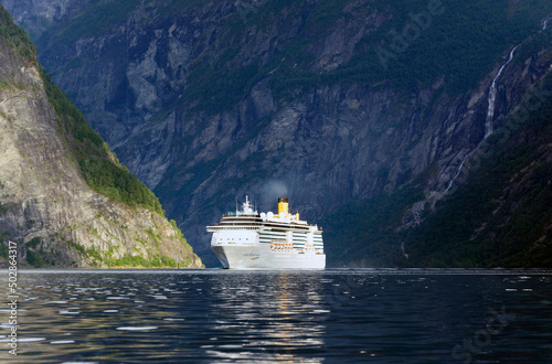 Cruise ship in the sea. Blue water of Geirangerfjord, dark blue and green Norwegian mountains, rays of sunshine on the ship and rocky slope